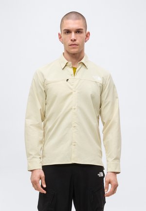 Νεαρός άνδρας με κοντά μαλλιά, φοράει μπεζ πουκάμισο με κουμπιά και μαύρο παντελόνι cargo με λογότυπα The North Face και στα δύο ρούχα.