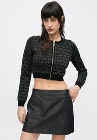 Tânără cu păr lung și închis la culoare, purtând un pulover negru scurt cu logo-ul MK repetat și o fustă mini gri închis, pozând cu mâna pe șold.