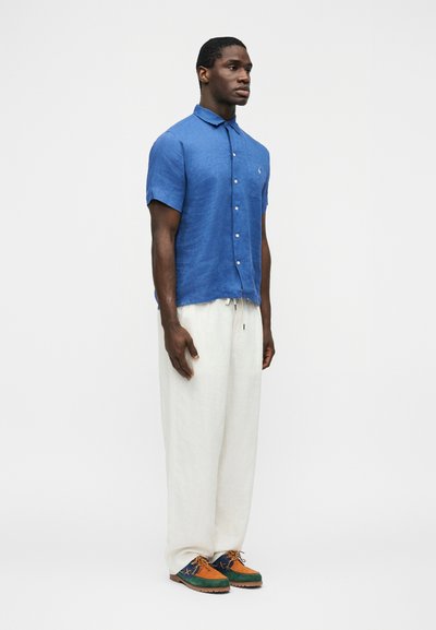 Vīrietis stāv valkājot zilu īsroku pogājamu krekliņu, vaļīgas baltas bikses un daudzkrāsainas zaļas, oranžas un zilas kurpes uz vienveidīga fona.