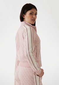 Veste zippée rose avec un motif texturé, accents rayés blancs et rose clair sur les manches, et poches latérales. Tissu doux avec une coupe ajustée.