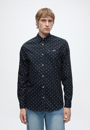 Junger Mann mit hellem Haar, der ein schwarzes Hemd mit weißen Diamantmustern und hellblaue Jeans trägt, steht vor einem schlichten weißen Hintergrund.