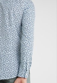 Camicia a maniche lunghe in cotone azzurro chiaro con un motivo floreale dettagliato in blu navy e verde. Presenta un polsino con bottone e un orlo curvato.