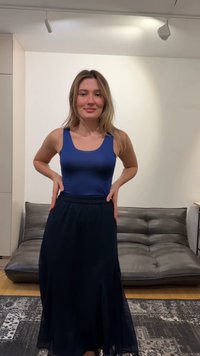 Fiatal nő áll fedett helyen, kezét csípőjén tartva, ujjatlan kék felsőt és hosszú sötét szoknyát visel, mögötte szürke kanapé és mintás szőnyeg látható.