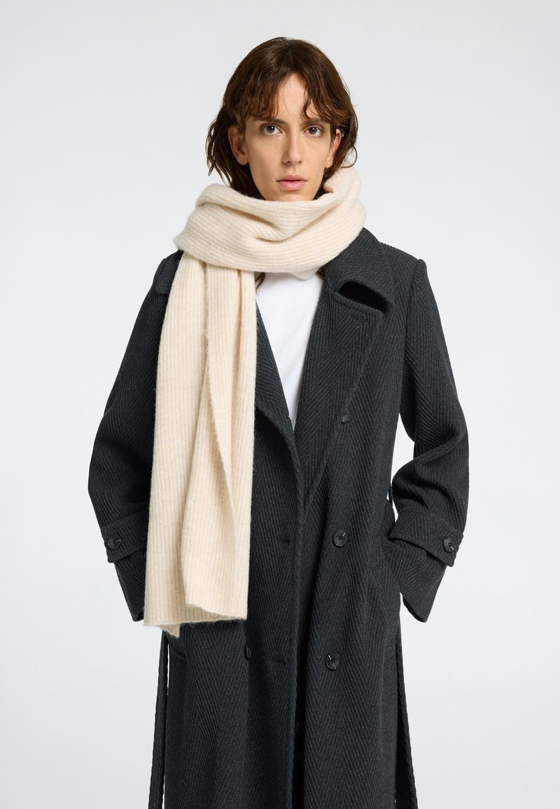 Beige ribbad scarf hängande runt nacken, i kombination med en mörk texturerad kappa med stora svarta knappar och en krage med snitt.