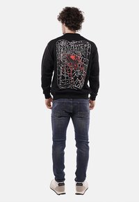 Felpa nera con un grande grafico di un ragno rosso e un intricato design di ragnatela bianca sul retro. Abbinata a jeans in denim blu.
