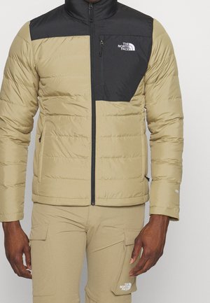 Homme portant une doudoune beige et noire North Face et un pantalon cargo beige avec des détails de fermeture éclair et de poches, montré du cou aux cuisses.
