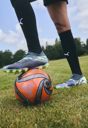 Botas de fútbol azules y verdes sobre un balón de fútbol con un diseño naranja y negro, situadas en un terreno de hierba, con patrones detallados.