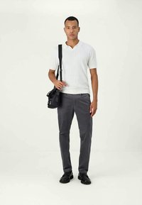 Man wearing white short-sleeve knit polo, gray pants, black shoes, and carrying a black shoulder bag on a plain background.