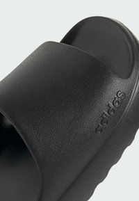 Black slide sandal with a textured finish and slightly raised footbed. Features a prominent embossed logo and smooth sole with wavy edge.