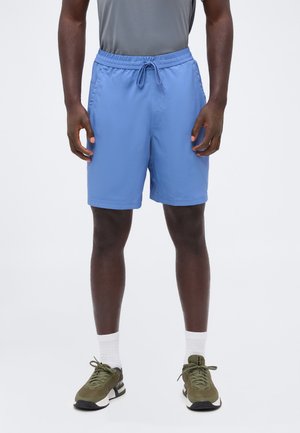 Homme portant un short bleu clair à cordon, un t-shirt gris, des chaussettes blanches et des chaussures de sport vert olive, debout sur fond blanc.