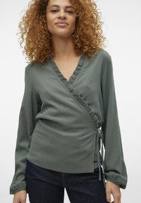 Haut vert à manches longues avec encolure à volants et détail de lien à la taille, fabriqué en tissu texturé. Porté avec un jean foncé.