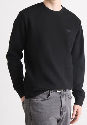 Man med svart långärmad sweatshirt med "BOSS"-logotyp och grå jeans, händer avslappnat vid sidorna mot en enkel bakgrund.