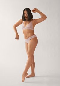 Lichtroze bralette en bijpassende slips gemaakt van zacht materiaal; de bralette heeft een driehoekig ontwerp en dunne bandjes. Model poseerde gracieus.