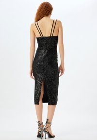 Robe en sequins noirs avec un design ajusté, longueur au genou et une fente dans le dos. Elle possède des fines bretelles et une surface texturée.