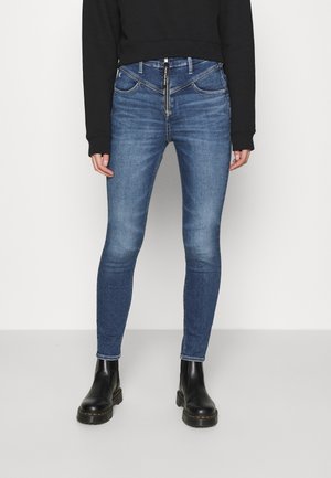 Person iført blå skinny jeans med frontlynlås og sorte ankelstøvler, kombineret med en sort langærmet top.