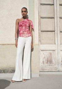 Rosa, einseitig geschnittenes Oberteil mit floralem Muster, kombiniert mit weißen, ausgestellten Hosen. Das Modell trägt silbernen Schmuck und weiße Sandalen. Hintergrund: abgenutzte Türen.