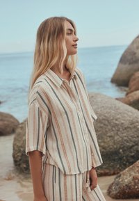 Young woman with long blonde hair wearing a striped button-up shirt and matching pants standing by large rocks near the sea.