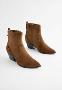 Botines de ante marrón hasta el tobillo con puntera puntiaguda, cremallera lateral y tacón apilado. Textura suave sin patrones o acentos visibles.