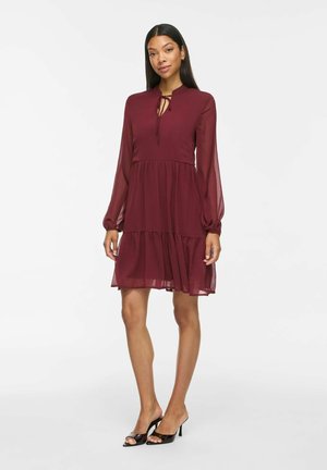 Femme debout portant une robe bordeaux à manches longues avec une jupe à volants et un col noué, associée à des chaussures à talons hauts noires à bout ouvert.