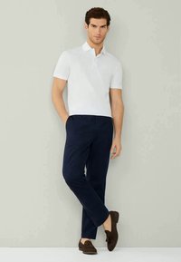 Witte poloshirt met korte mouwen, gecombineerd met marineblauwe broek. Model staat casual, met donkere bruin leren instappers. Eenvoudige, moderne outfit.