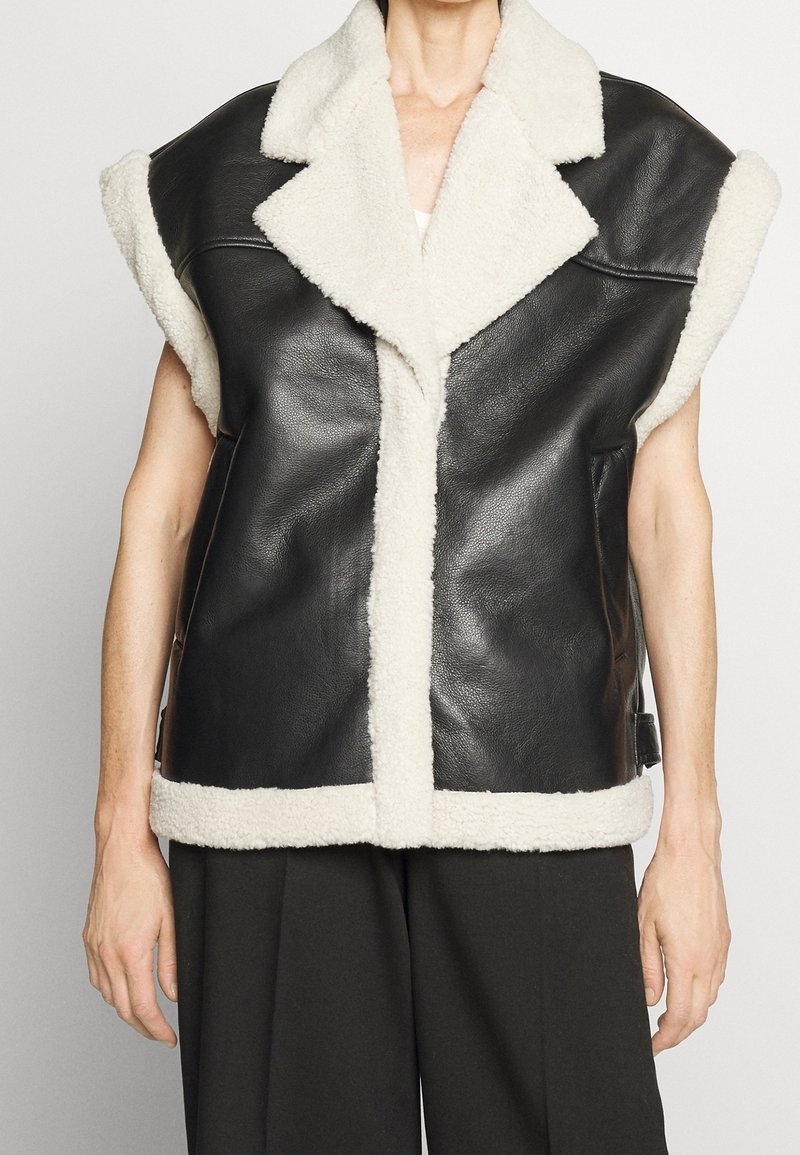 Gilet en cuir noir sans manches avec doublure en shearling blanc sur le col, les bords avant, les poignets et l'ourlet porté sur un pantalon noir.