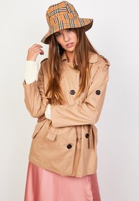 Gabardina beige con botones negros, combinada con un sombrero tipo bucket de cuadros. El conjunto incluye una falda rosa brillante de satén y puños de suéter blancos acanalados.