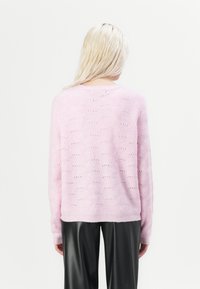 Een persoon met lang, steil blond haar draagt een lichtroze sweater en zwarte leren broeken, met de rug naar ons gekeerd tegen een eenduidige witte achtergrond.