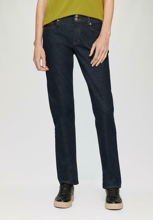 Jean en denim bleu foncé à coupe droite, avec des coutures orange et du matériel en métal, porté avec des baskets noires. Design minimaliste.