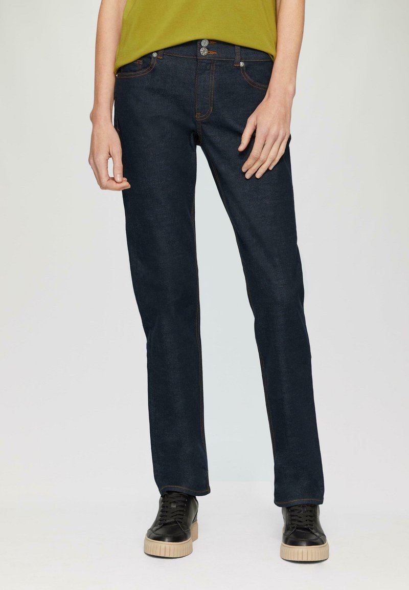 Dunkelblaue Jeans im geraden Schnitt, mit orangefarbenen Nähten und Metallbeschlägen, getragen mit schwarzen Sneakers. Minimalistisches Design.