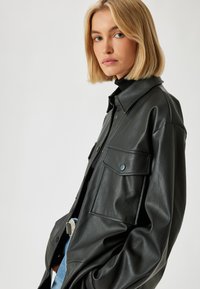 Schwarze Lederjacke mit kragenförmigem Hals, zwei Fronttaschen und lockerer Passform. Die Textur ist glatt, mit sichtbaren Nähdetails.