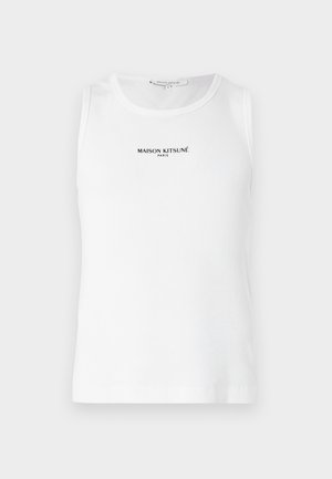 Giovane uomo con capelli ricci che indossa una canotta bianca, pantaloni neri larghi e scarpe da ginnastica bianche, in piedi con le mani nelle tasche.