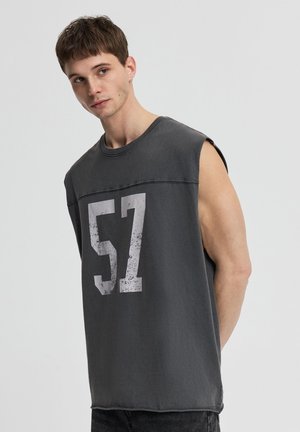 Giovane uomo che indossa una maglietta senza maniche grigio scuro con una grande stampa consumata "57", guarda di lato contro uno sfondo semplice.