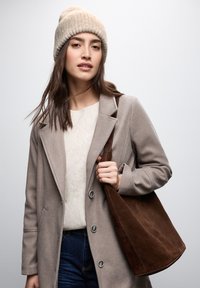 Cappotto in misto lana beige con rever sagomati, maglione a maglia color crema e borsa a tracolla in suede marrone. Il modello indossa un berretto di maglia chiaro.