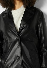 Czarna kurtka ze sztucznej skóry z klapą z wycięciem, zapinana na pojedynczy guzik, z bocznymi kieszeniami. Noszona na białym swetrze o fakturze.
