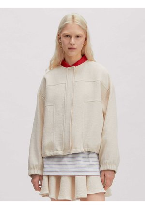 Blonde Frau mit goldenen Ohrringen, die eine strukturierte cremefarbene Jacke über einem roten Hemd mit Kragen und einem gestreiften Rock vor einem einfarbigen Hintergrund trägt.