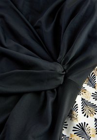 Crni materijal s detaljem u obliku uvrnute čvorke, uparen s bijelim materijalom s uzorkom koji sadrži zlatne i crne cvjetne dizajne. Glatka tekstura.
