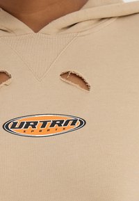 Béžová mikina s kapucí s žebrovanou texturou, s dvěma ošuntělými výřezy a oválným logem URTRN SPORTS v oranžové a černé barvě.