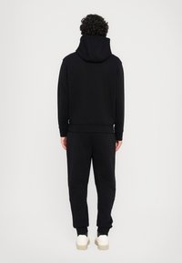 Schwarze Kapuzenjacke mit hohem Kragen, lockerer Passform und gerippten Bündchen, kombiniert mit passenden Jogginghosen und weißen Sneakers. Glatte Textur, minimalistisches Design.