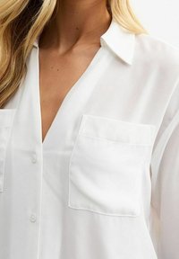 Blusa bianca, leggera, con colletto, bottoni frontali e due tasche sul petto. Texture liscia con vestibilità rilassata e materiale leggermente trasparente.