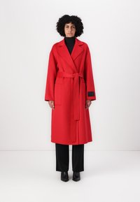Roter Wollmischmantel mit einem Bindegürtel, breiten Revers, großen Fronttaschen und langen Ärmeln, kombiniert mit schwarzen Hosen und Stiefeletten.