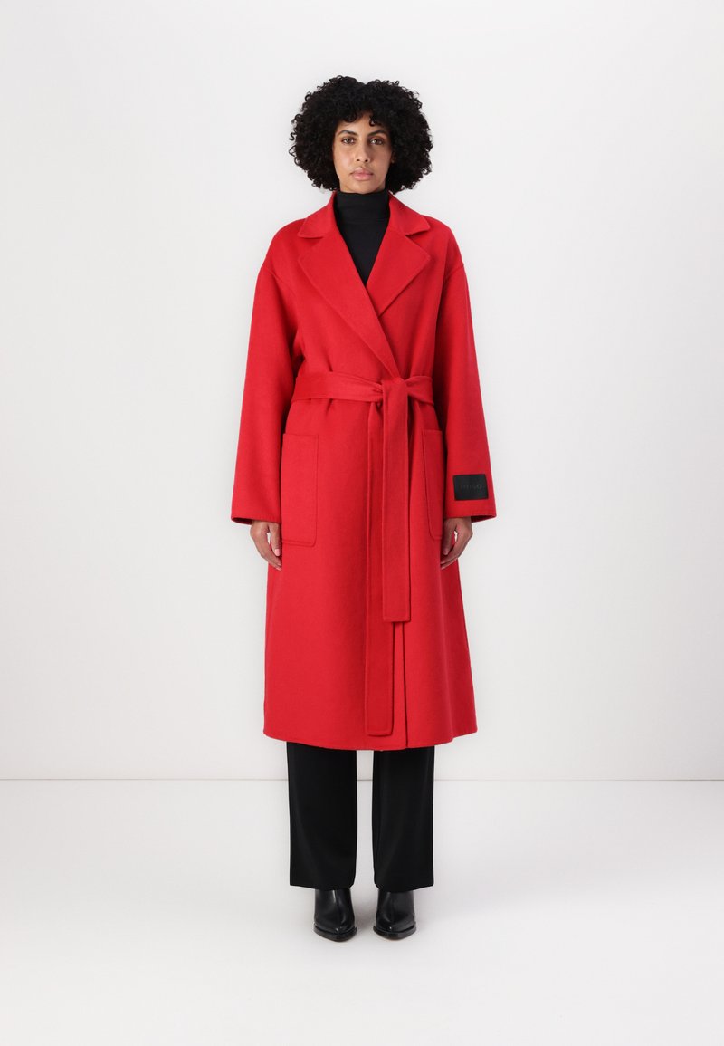 Roter Wollmischmantel mit einem Bindegürtel, breiten Revers, großen Fronttaschen und langen Ärmeln, kombiniert mit schwarzen Hosen und Stiefeletten.