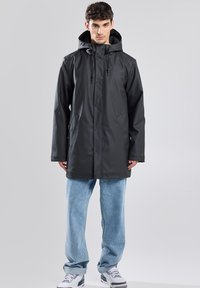 Impermeabile nero con cappuccio, chiusura anteriore a bottoni automatici e coulisse regolabili; abbinato a jeans azzurri larghi.
