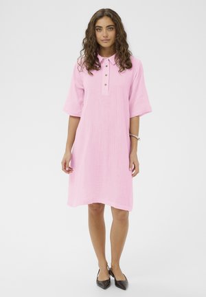 Woman with curly hair wearing a pink buttoned, knee-length dress and black pointed shoes, standing against a light background.