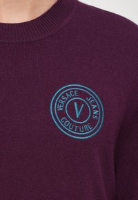 Vijolična pletena jopica z okroglo turkizno vezeno oznako, ki se glasi "VERSACE JEANS COUTURE." Teksturovani material z rebrastim izrezom.