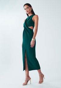 Zelena oprijeta obleka z zavitim korzetom in stranskimi izrezi, z visokim razporkom na stegno. Kombinirana z zlato obutvijo z naramnico in zapestnico.