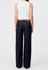 Femme portant un jean large bleu foncé avec une ceinture marron et un haut sans manches blanc, vue de dos sur un fond uni.