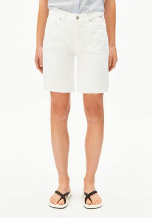 Femme portant un short en denim blanc taille haute avec ourlet brut, associé à des sandales slip-on noires et blanches, debout devant un fond blanc.