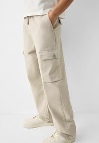 Beżowe spodnie cargo w luźnym kroju, z bocznymi kieszeniami, elastycznym paskiem i gładką teksturą z mieszanki bawełny. Noszone z białymi trampkami.