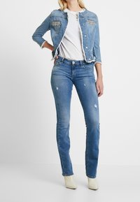 Chaqueta de mezclilla azul clara con bordes deshilachados, bolsillos adornados, combinada con jeans rectos azul claros que presentan detalles desgastados y calzado blanco.
