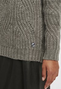 Primo piano di una mano appoggiata accanto a un maglione grigio a trecce, con un piccolo tag metallico rotondo e pantaloni neri di pelle.